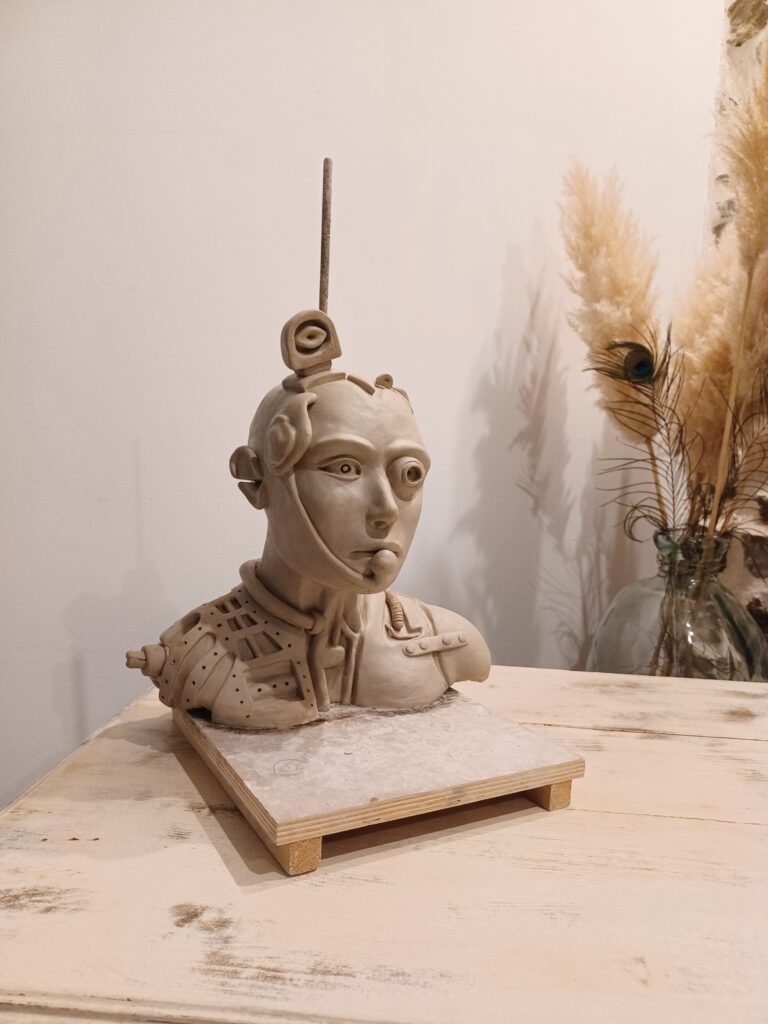 Sculpture de tête de cyborg en argile blanche sur tige et posée sur un socle de travail en bois, avant cuisson.